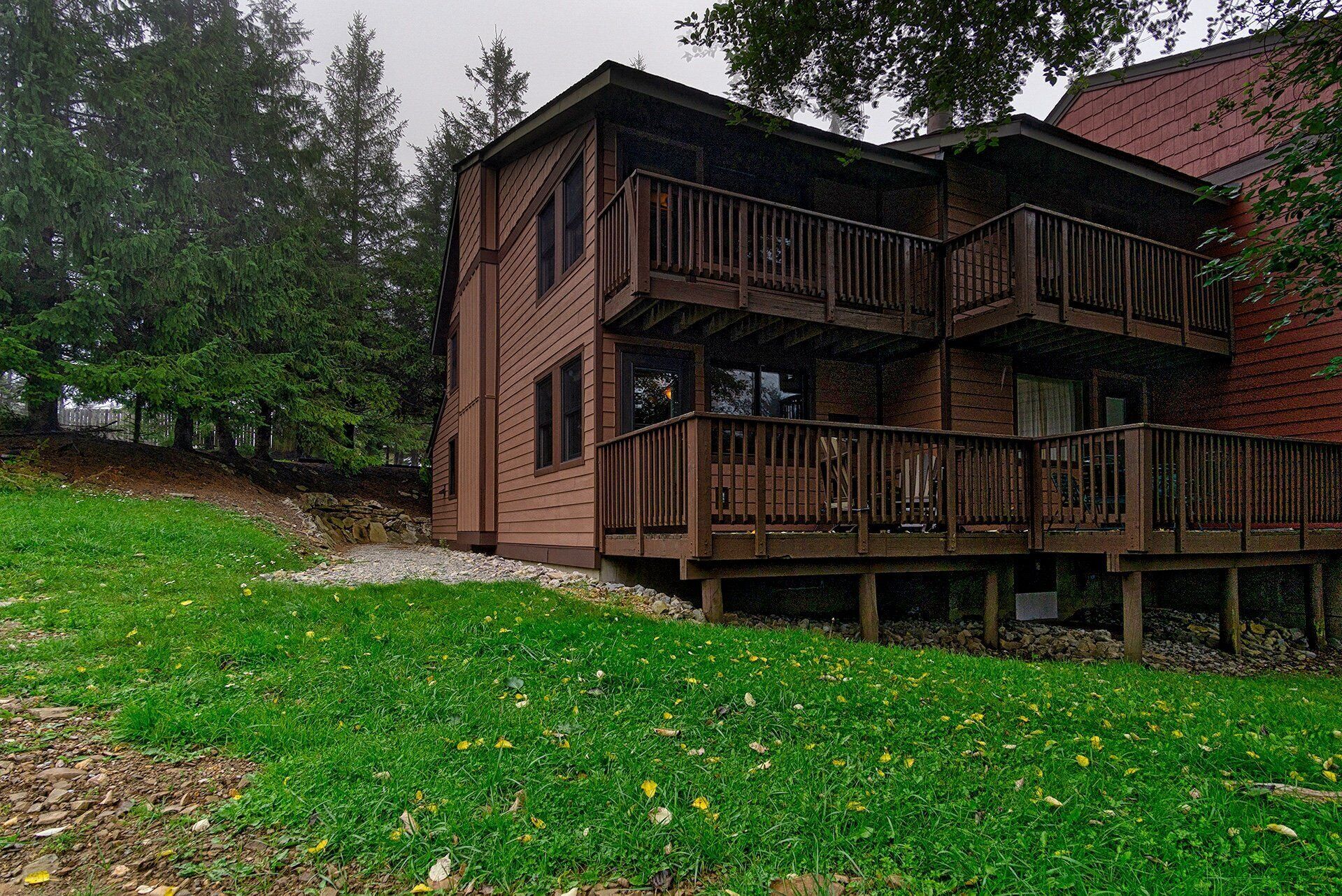 Uma cabana de madeira de dois andares com varanda, rodeada por gramado e árvores.