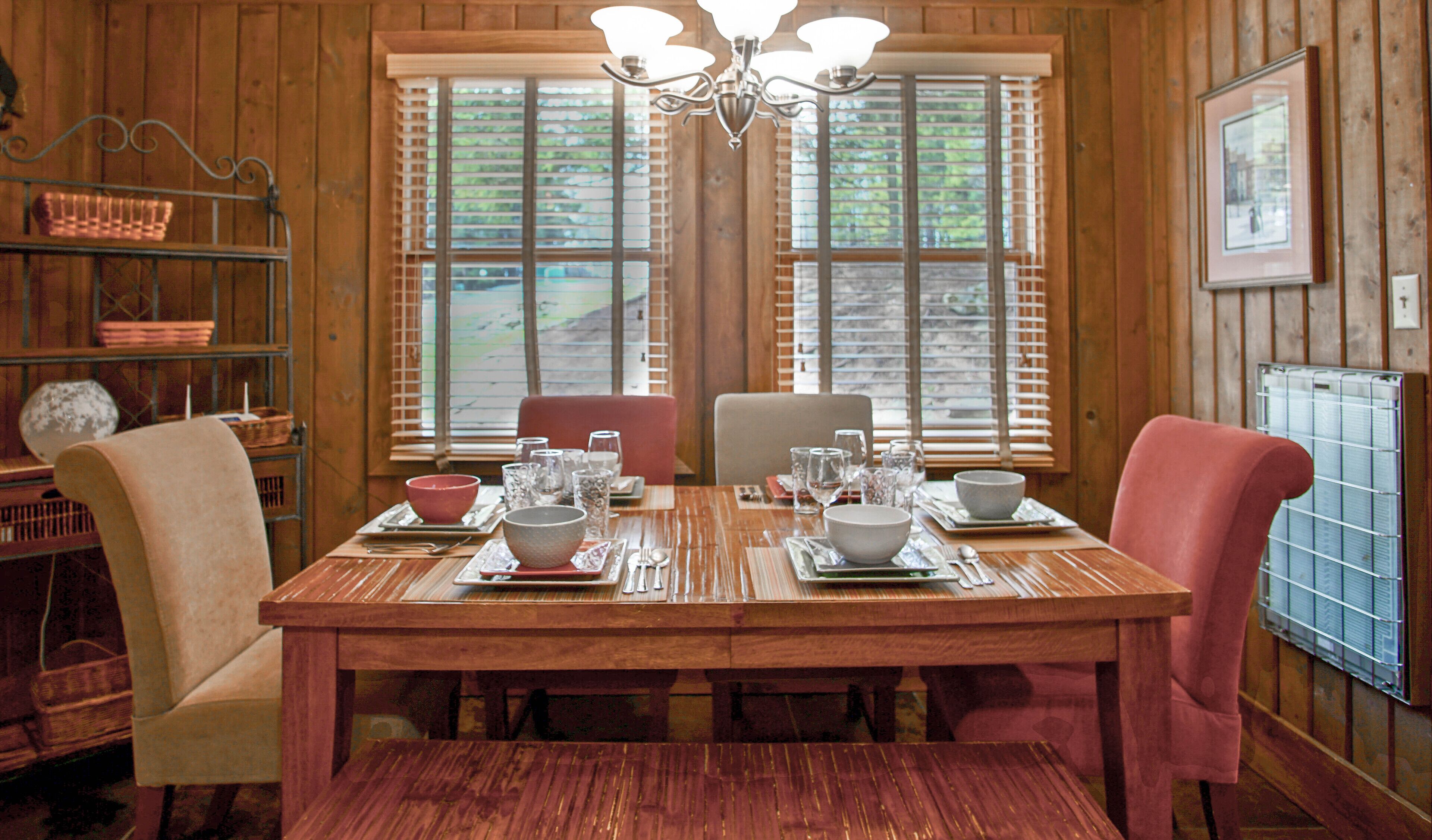 A wooden dining table set with bowls, glasses, and cutlery, flanked by chairs, in a room with wooden walls and a window with blinds.
