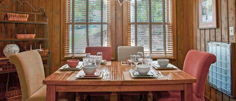 A wooden dining table set with bowls, glasses, and cutlery, flanked by chairs, in a room with wooden walls and a window with blinds.