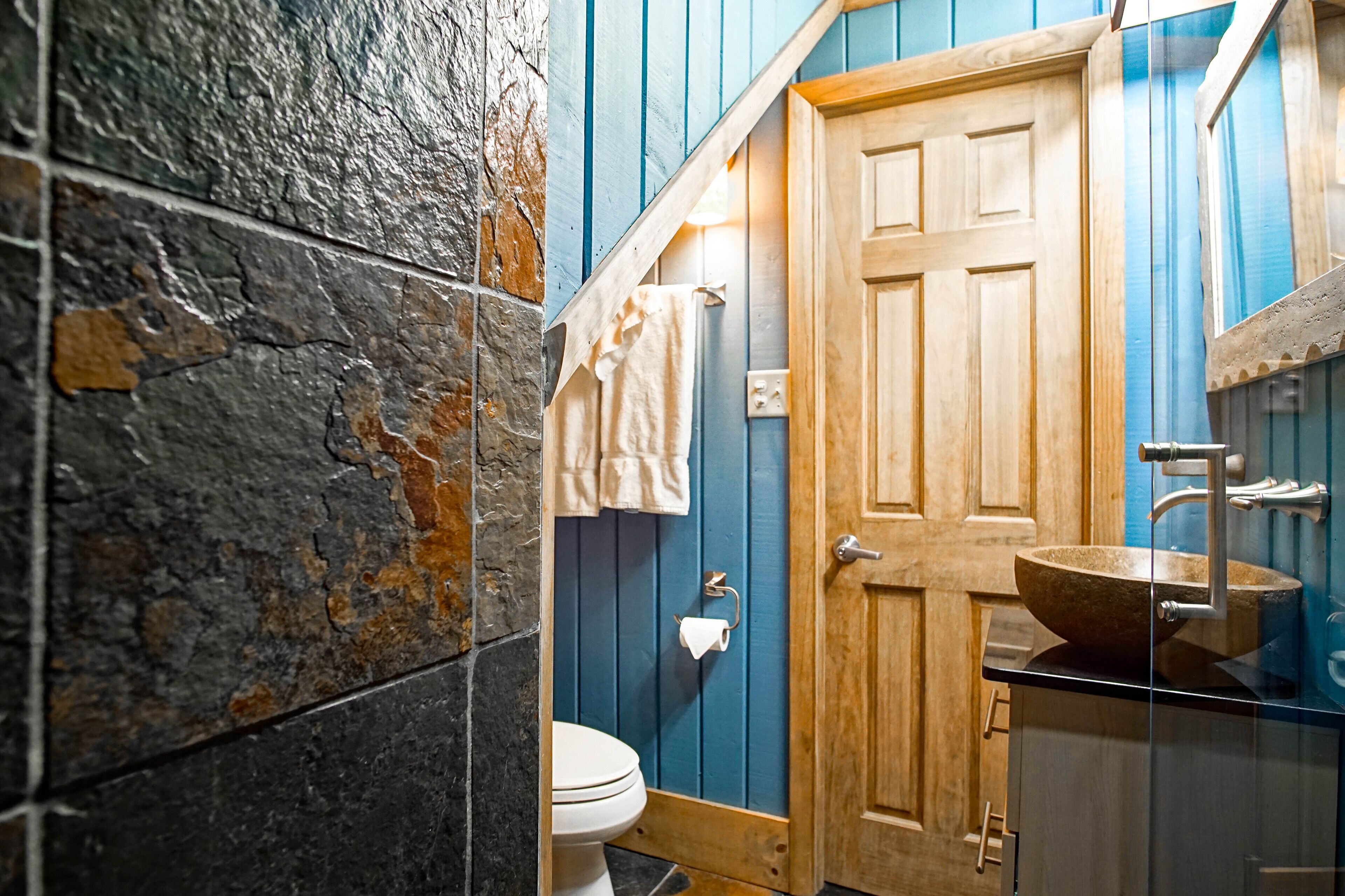 A bathroom with a stone wall, a wooden door, a white toilet, and a wooden vanity with a sink.