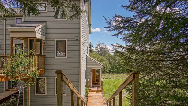 A two-story house with a wooden deck leading to a front door, surrounded by trees. - Powderidge 1 Hideaway Slope Side (Snowshoe)