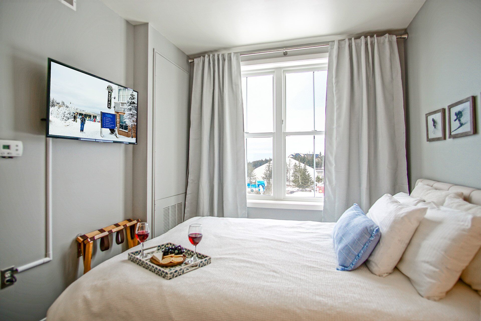 A hotel room with a bed, a flat-screen TV mounted on the wall, a window with curtains, and a tray with food and drinks on the bed.