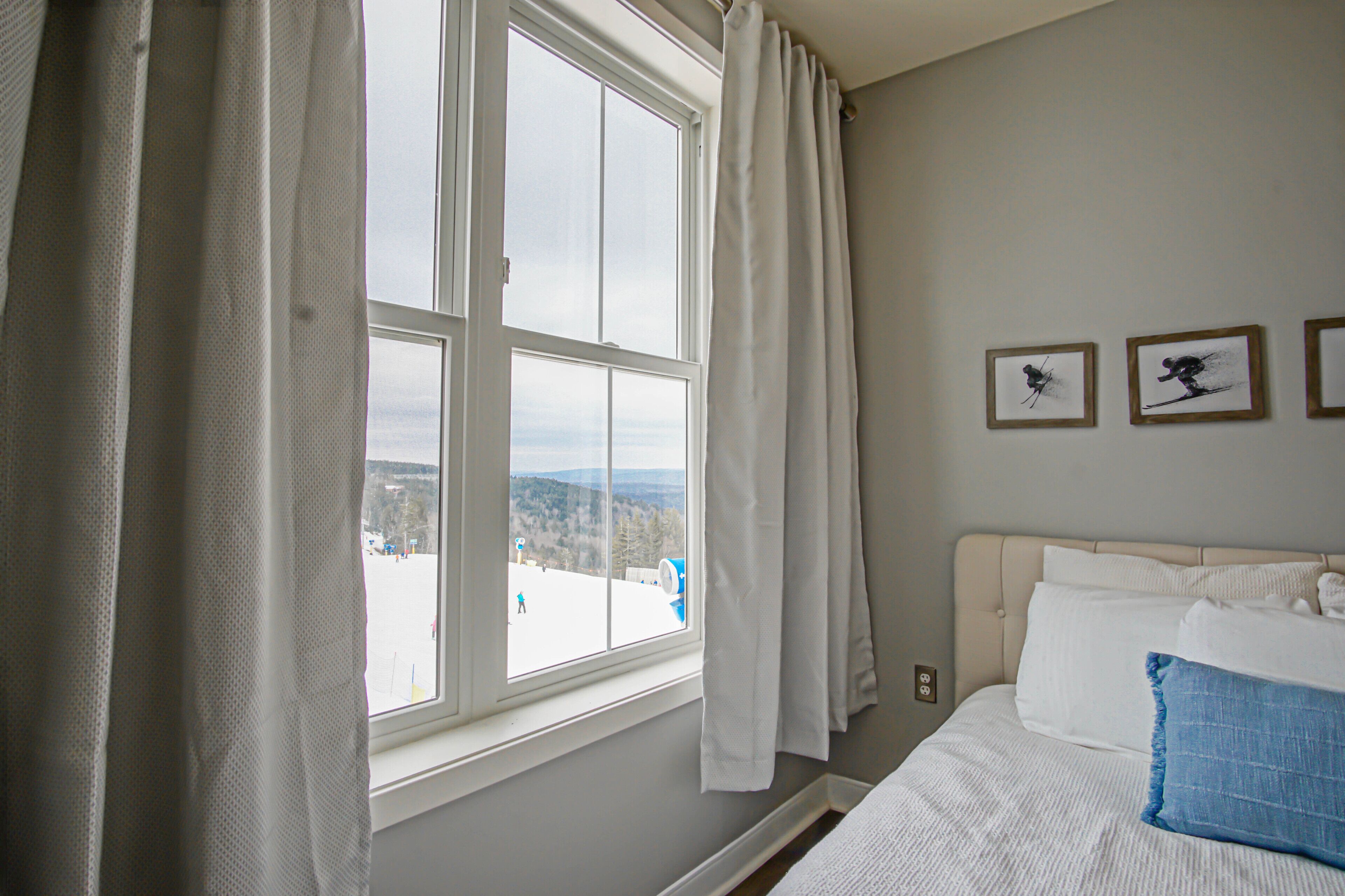 A bedroom with a large window, a bed with white bedding, and two framed pictures on the wall.