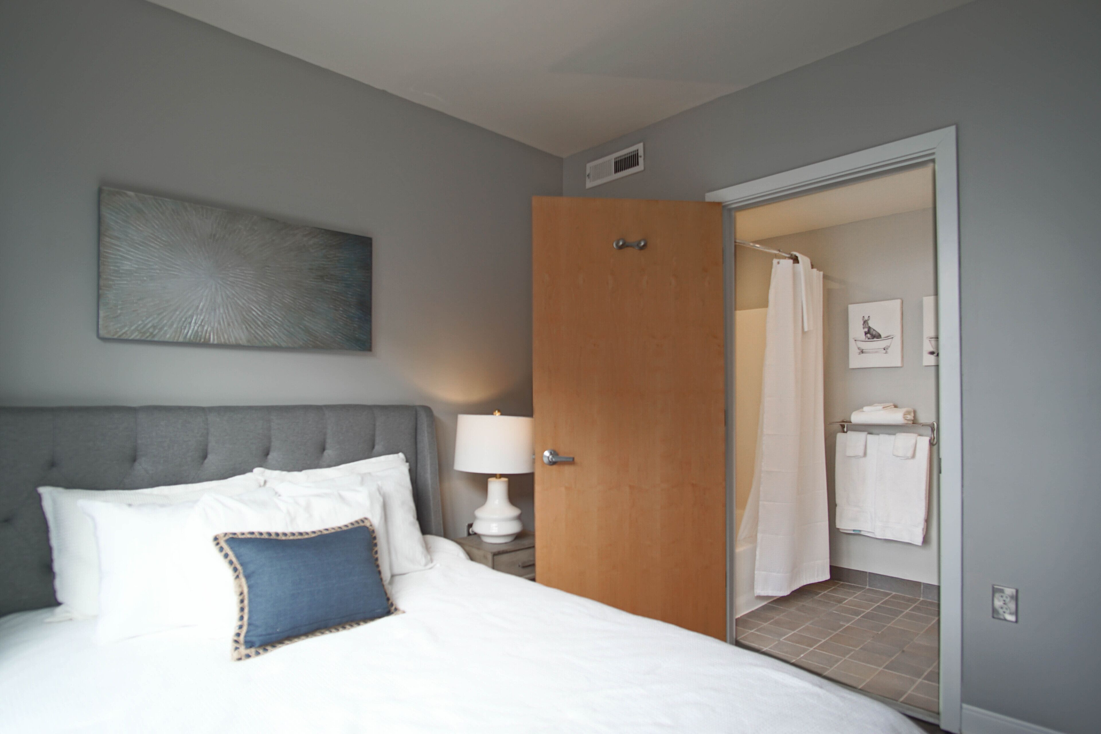Una habitación de hotel con una cama, una mesita de noche con lámpara, una puerta de madera que da a un baño y una obra de arte colgada en la pared.