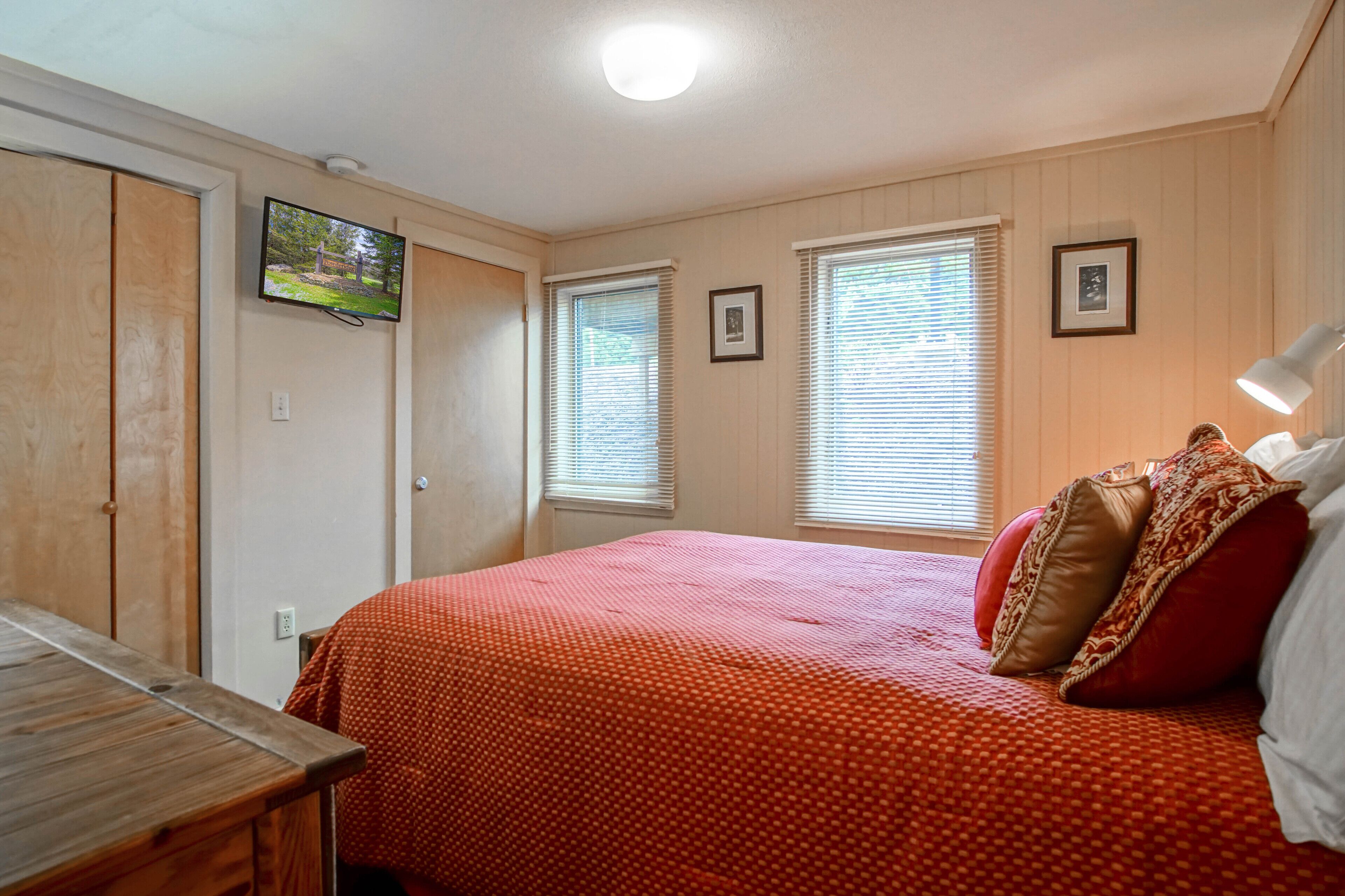 A bedroom with a wooden bed, a TV mounted on the wall, and two windows with blinds.