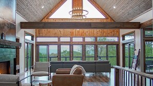 A spacious living room with a high ceiling, wooden beams, a fireplace, and large windows offering a view of the outdoors. - 5 Br: Cupp Runneth Over - Luxurious Retreat Nestled 9.7 Miles From Snowshoe (Slaty Fork)