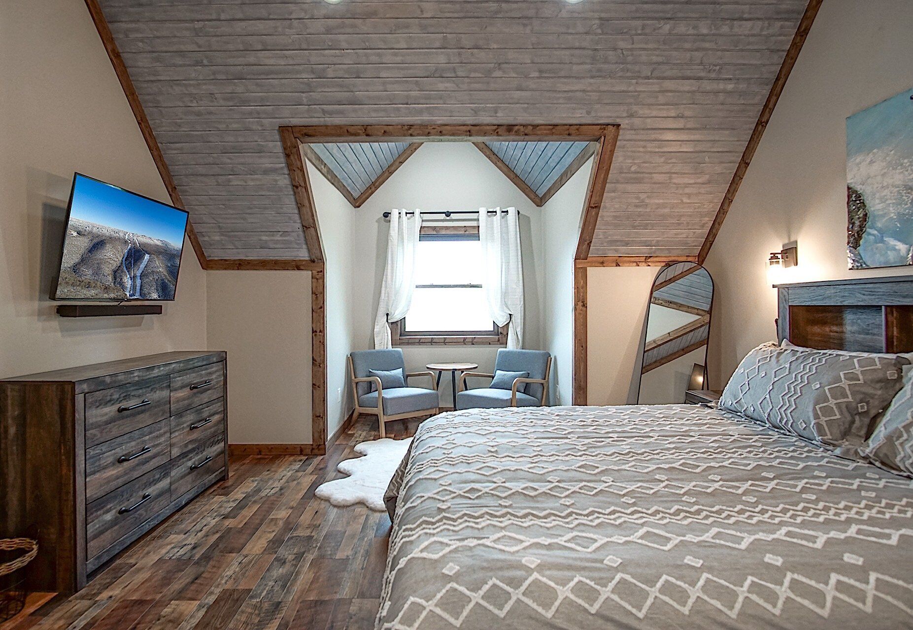 Un dormitorio con techo inclinado, una cama con ropa de cama estampada, un aparador, un televisor y una ventana con cortinas.