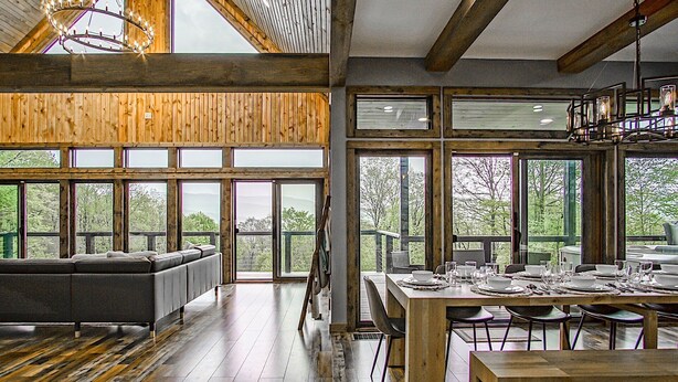 A spacious dining area with a wooden table set for a meal, large windows offering a view of the outdoors, and a cozy seating area with a sofa. - 5 Br: Cupp Runneth Over - Luxurious Retreat Nestled 9.7 Miles From Snowshoe (Slaty Fork)