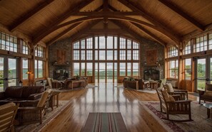 Amplia habitación con techos altos y ventanas grandes, que incluye chimenea, muebles de madera y vistas al exterior.