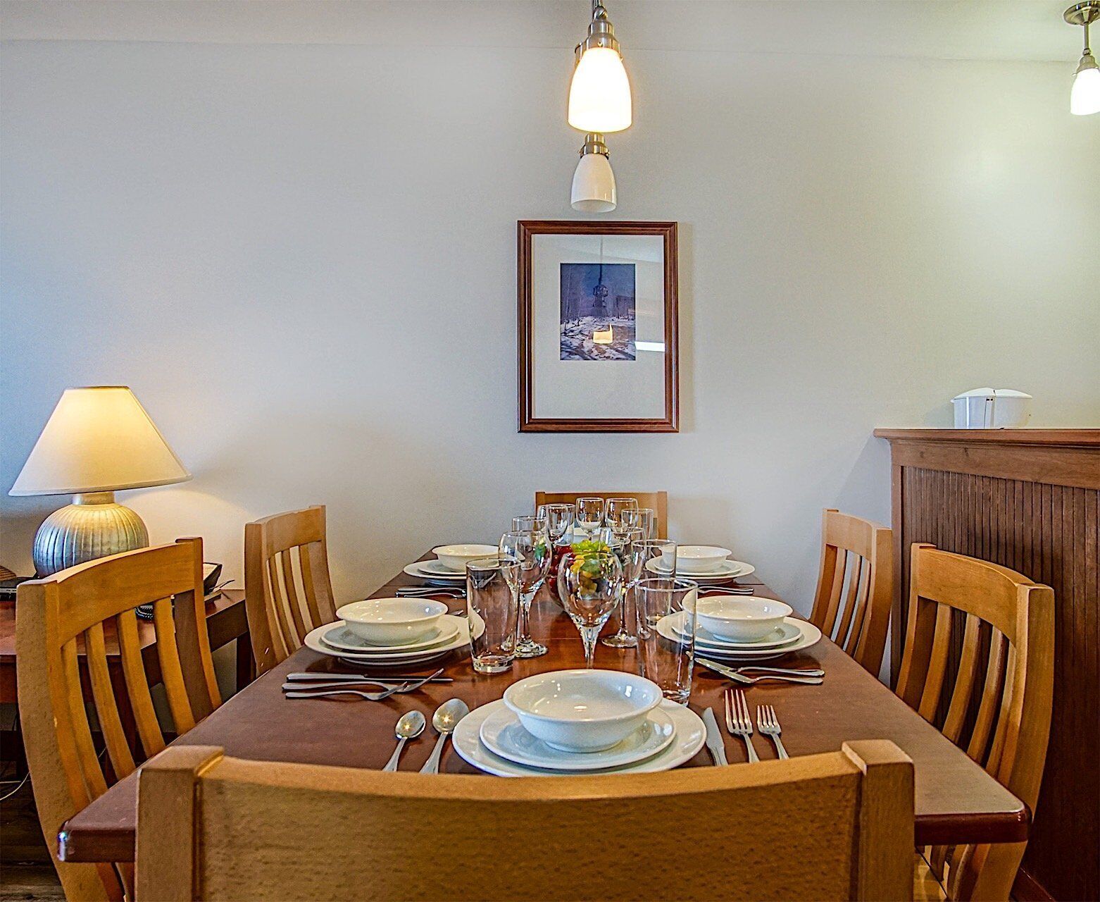 A dining area with a wooden table set for four, surrounded by wooden chairs. There is a framed picture on the wall above the table.