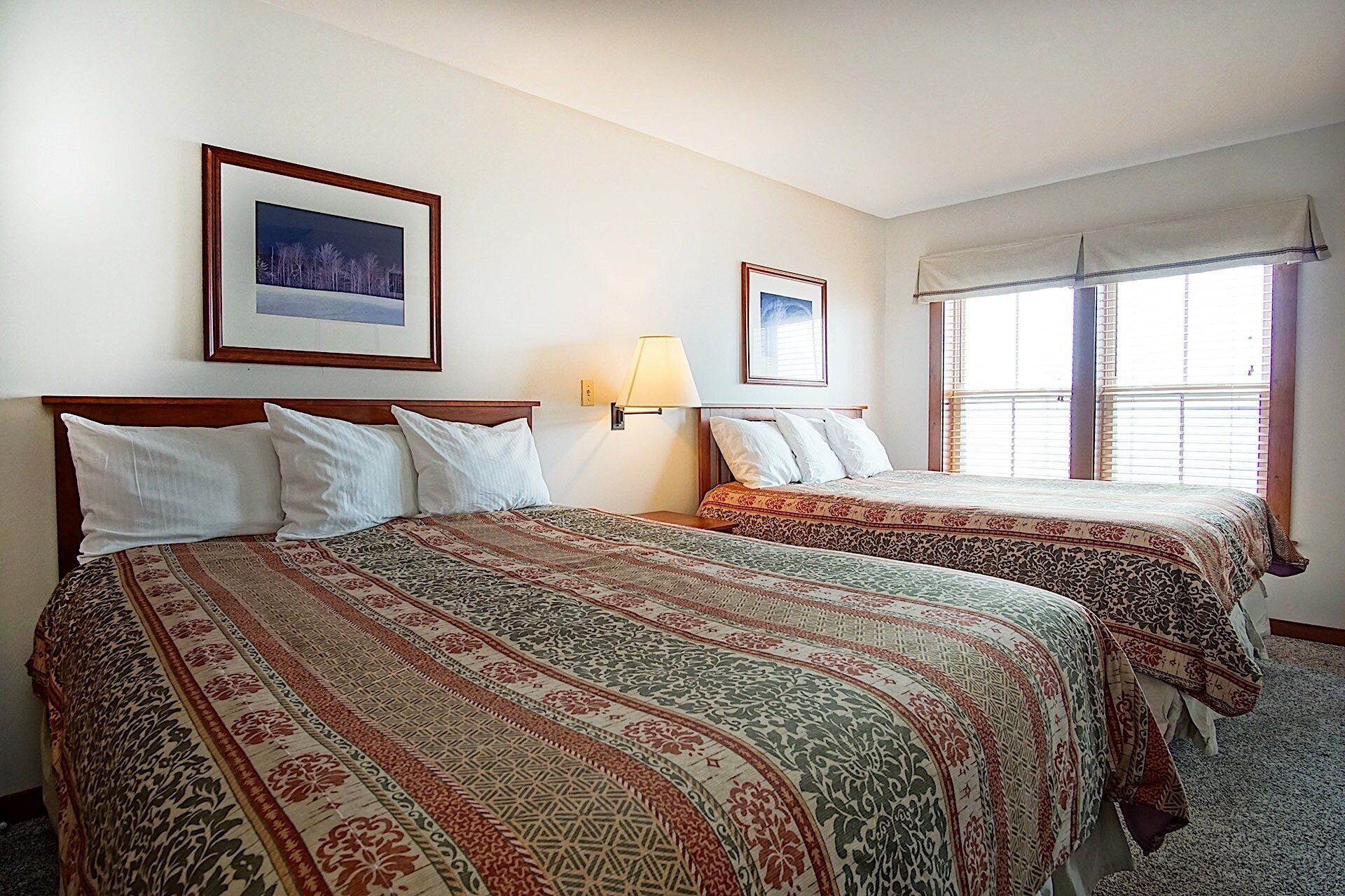 A hotel room with two beds, each with a patterned bedspread, a nightstand with a lamp, and a window with blinds.
