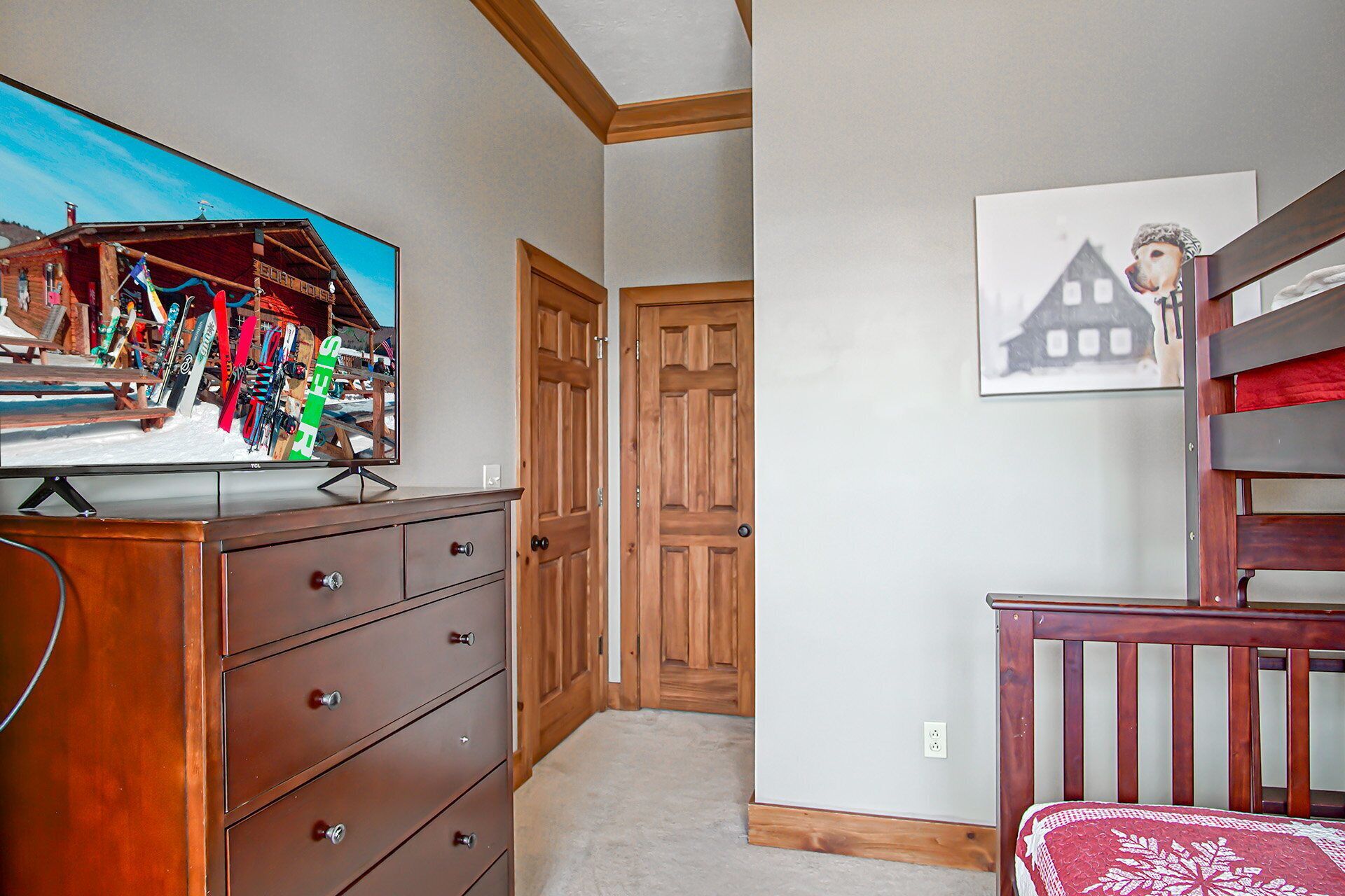 A room with a wooden dresser, a television displaying a ski resort, a wooden door, and a crib with a red blanket.