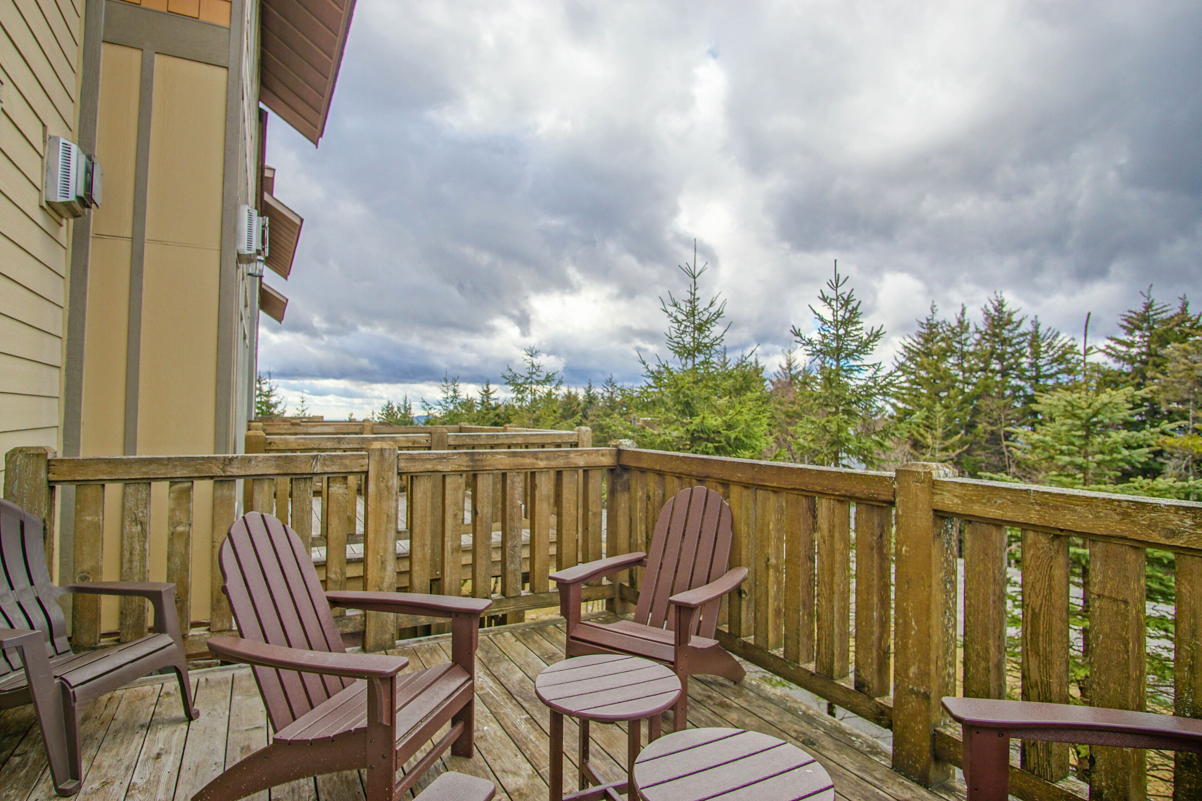Eine Holzterrasse mit Adirondack-Stühlen und einem Tisch, mit Blick auf ein bewaldetes Gebiet unter einem bewölkten Himmel.