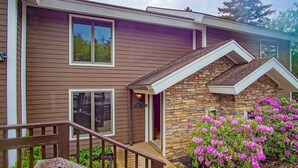 Une maison à deux étages avec une façade en pierre, une terrasse en bois et un arbuste à fleurs roses dans le jardin.