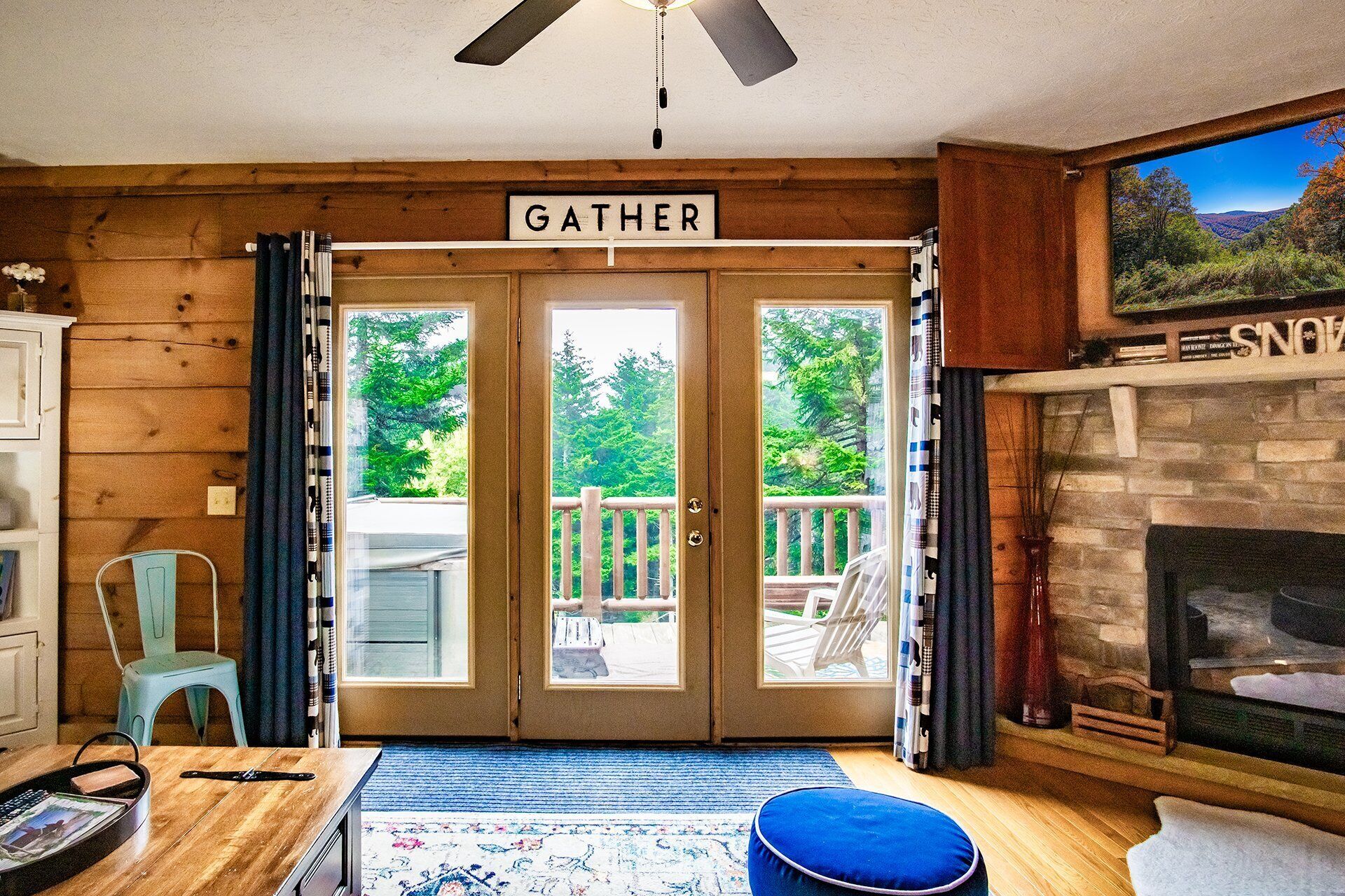 A cozy living room with a fireplace, a 'Gather' sign above the double doors, and a view of the outdoors through the doors.