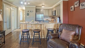A kitchen with a central island, bar stools, a leather chair, and wooden cabinets. - Powderidge 44: 1br: 1 Bath Flat (Snowshoe)