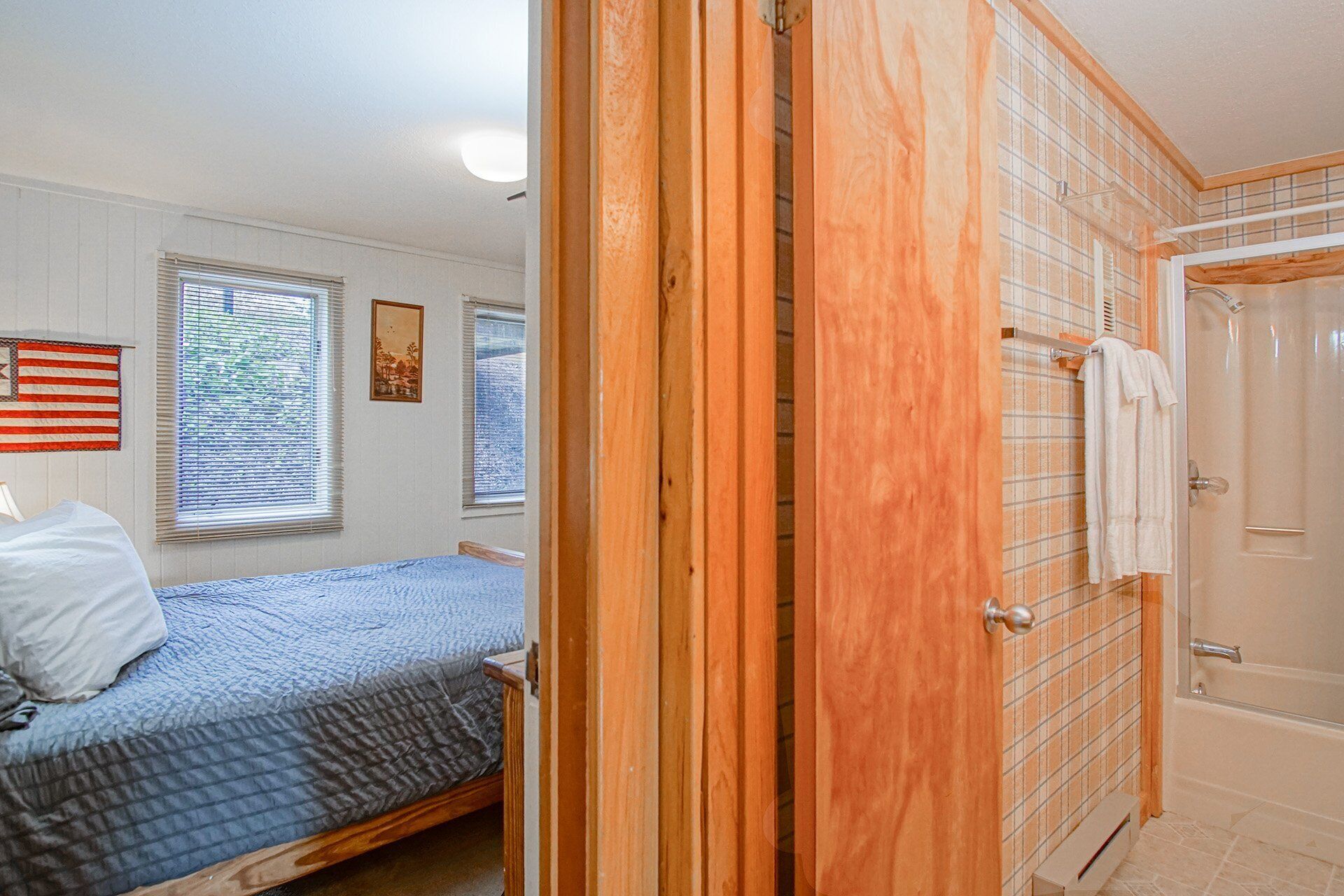 Ein Schlafzimmer mit einem Bett, einem Fenster mit Jalousien, einem gerahmten Bild und einer Tür zu einem Badezimmer mit Dusche und Handtuchhalter.