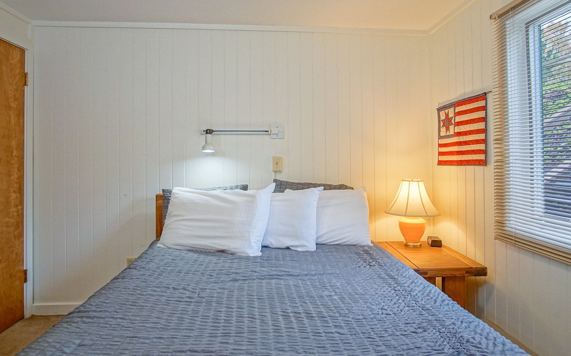A bedroom with a bed, a nightstand, a lamp, a window with blinds, and a wall with a quilt.