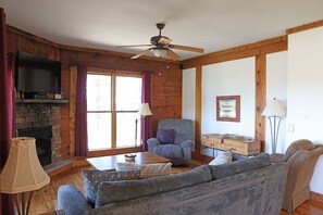 A cozy living room with a stone fireplace, wooden walls, a ceiling fan, a television, a sofa, a chair, and a lamp. - 4 BR: Timbers #1 Stand Alone Cabin (Snowshoe)