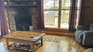 A cozy living room with a stone fireplace, wooden flooring, and a large window with curtains. - 4 BR: Timbers #1 Stand Alone Cabin (Snowshoe)