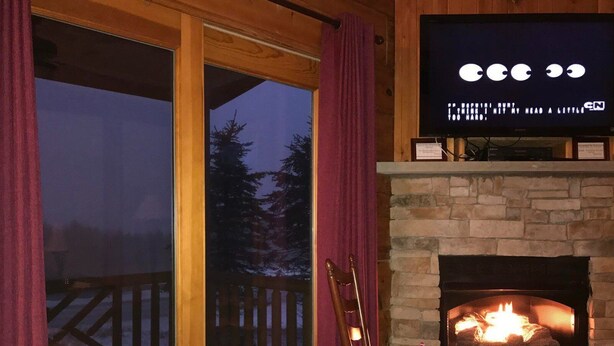 A cozy room with a fireplace, a television mounted above it, and a large window with curtains. - 4 BR: Timbers #1 Stand Alone Cabin (Snowshoe)