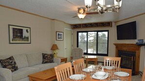 Une salle à manger avec une table en bois dressée pour six personnes, un canapé, une cheminée et un ventilateur de plafond.