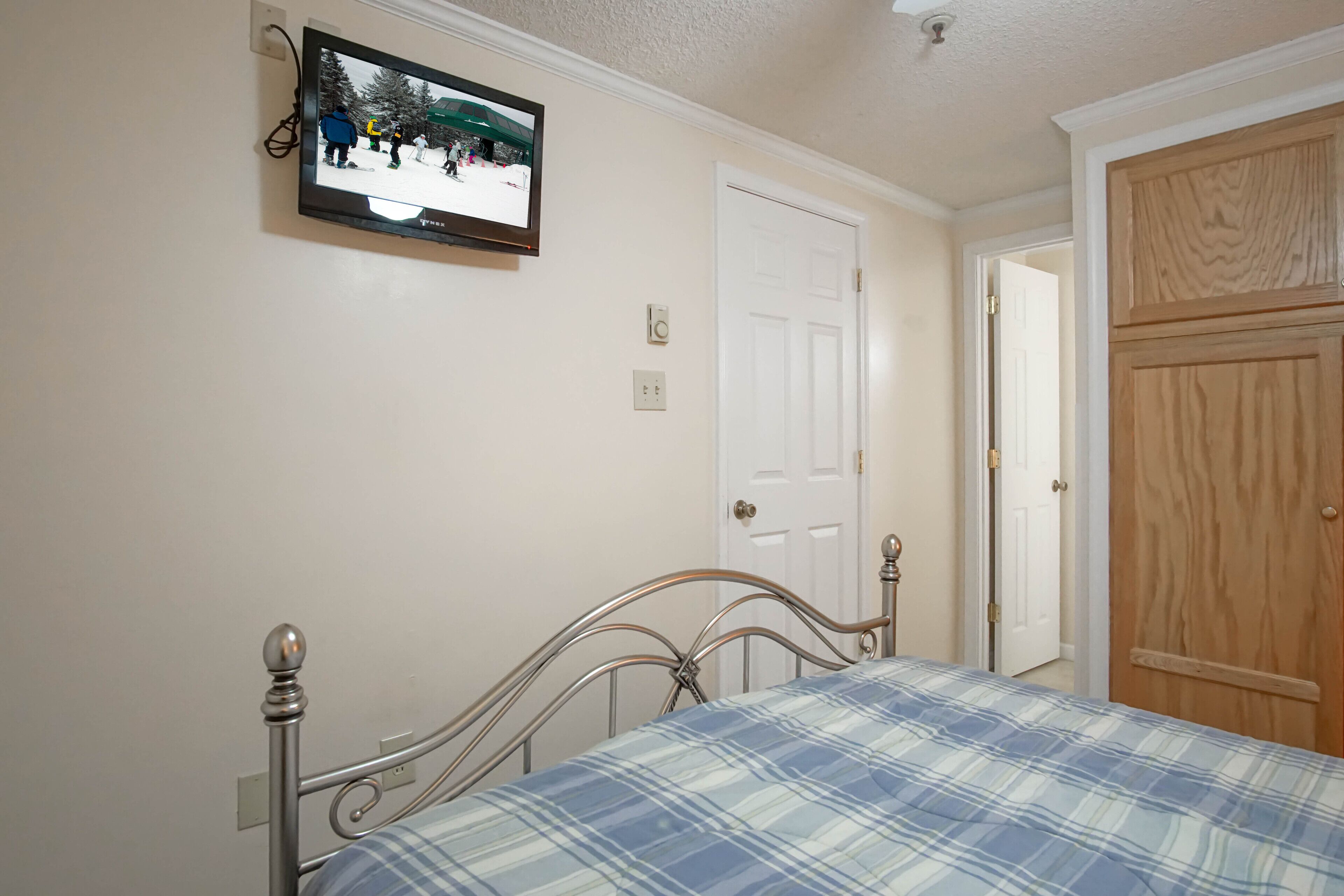 A bedroom with a metal bed frame, a plaid bedspread, a television mounted on the wall, and a wooden door.