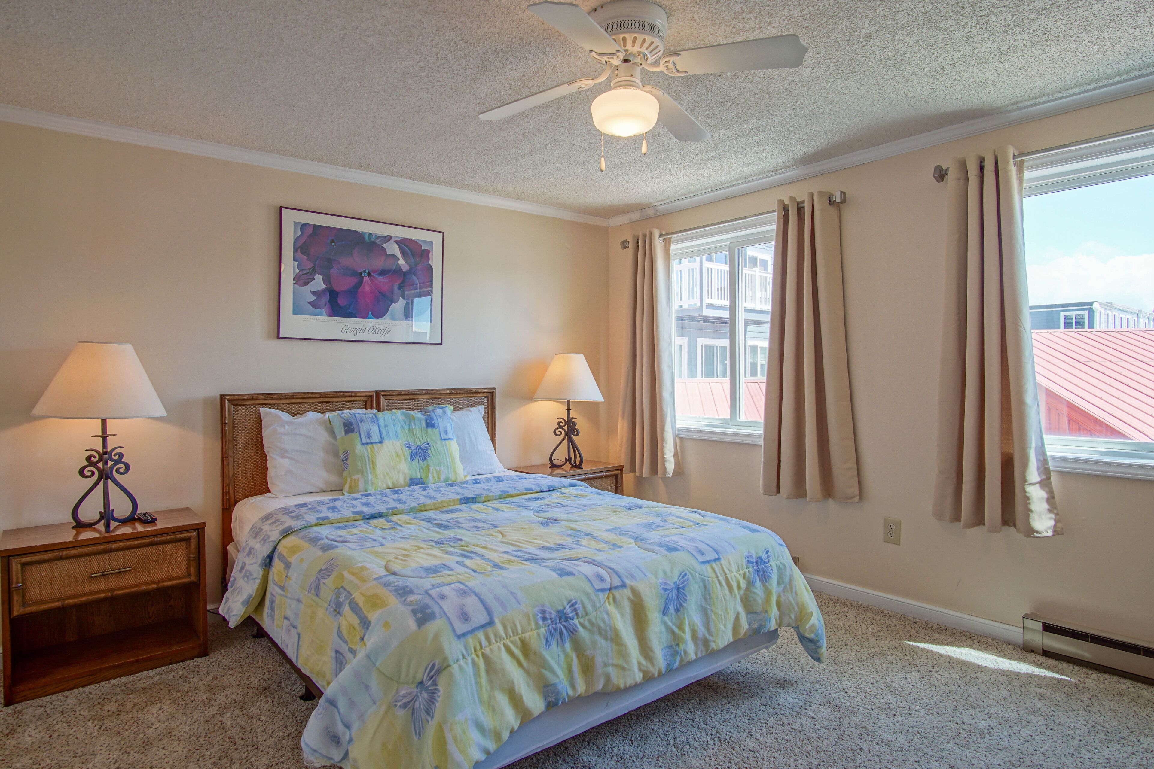 A bedroom with a bed, two bedside lamps, a ceiling fan, a window with curtains, and a framed picture on the wall.