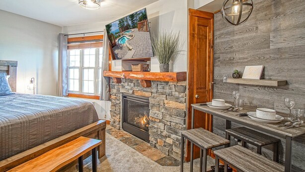 A bedroom with a stone fireplace, a bed with a view of the outdoors, a dining area with a table and benches, and a hanging light fixture. - Rimfire 421 Cozy Studio Just Steps From the Village Slope Side (Snowshoe)
