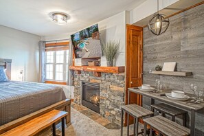 A bedroom with a stone fireplace, a bed with a view of the outdoors, a dining area with a table and benches, and a hanging light fixture. - Rimfire 421 Cozy Studio Just Steps From the Village Slope Side (Snowshoe)