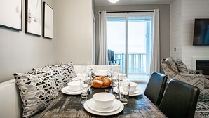 Un coin repas avec une table dressée pour quatre personnes, un canapé agrémenté de coussins à motifs et une vue sur l’extérieur grâce à une grande fenêtre.