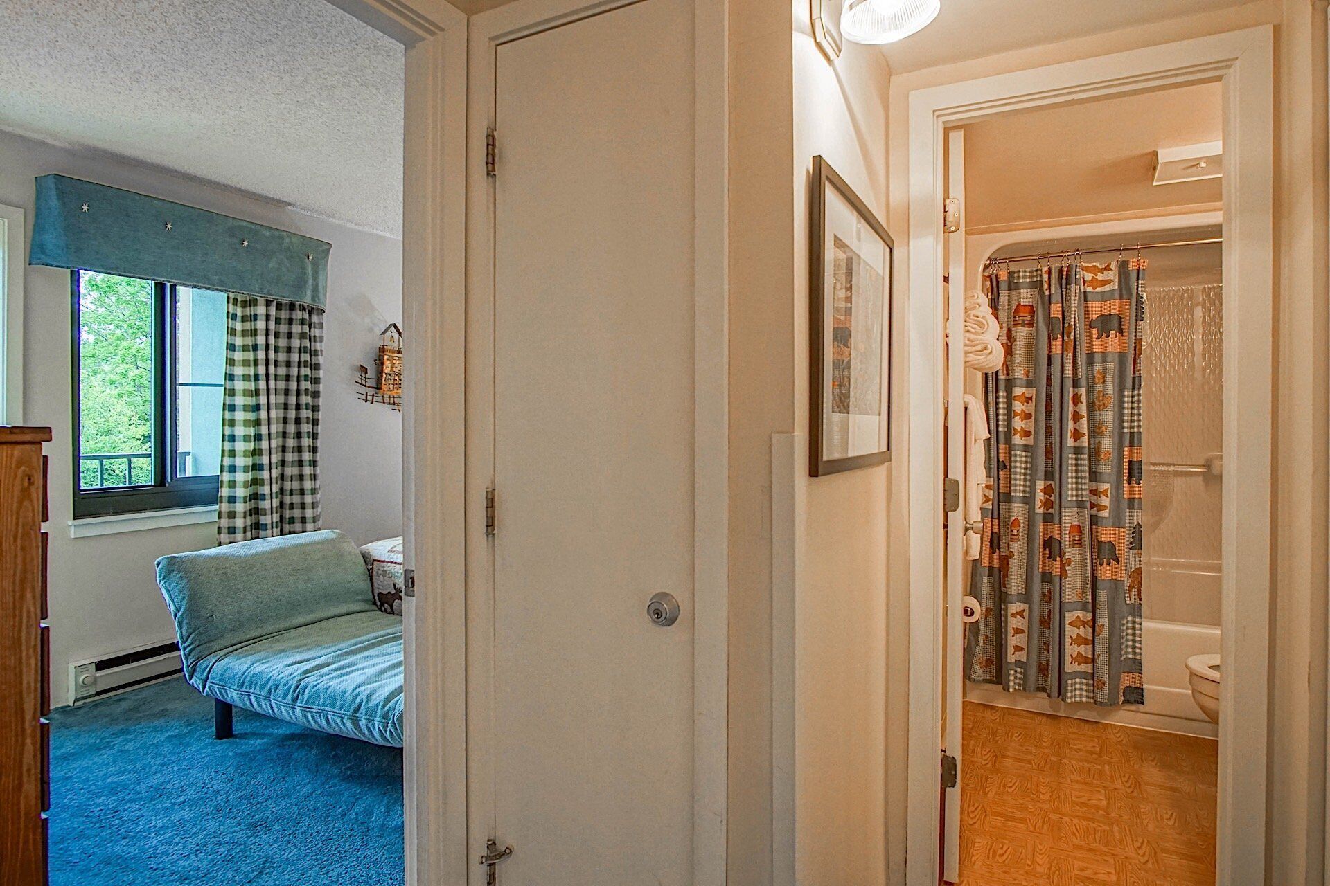 A bedroom with a bed, a window with curtains, a radiator, and a door leading to a bathroom with a shower curtain.