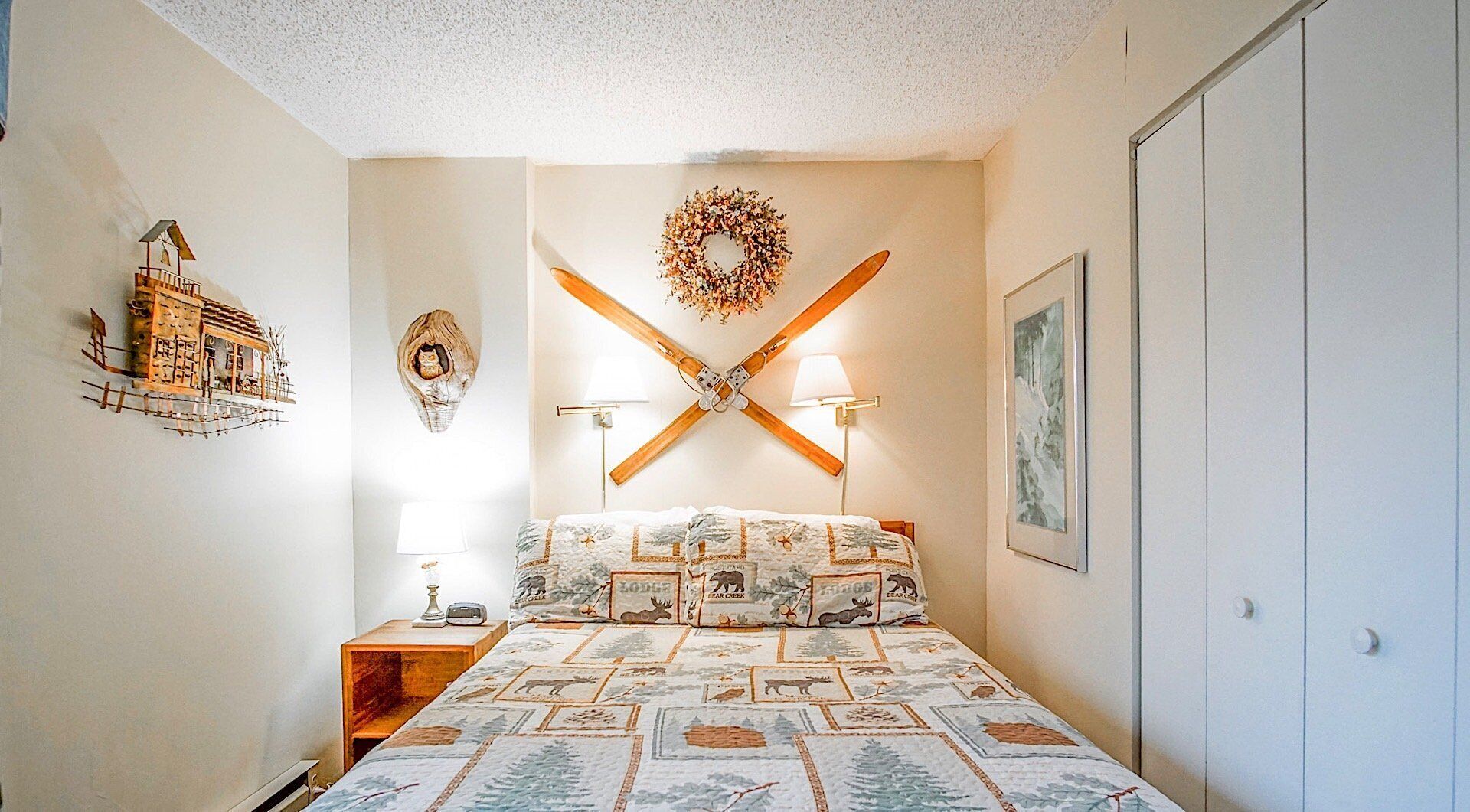 A bedroom with a bed, a nightstand, a wall-mounted lamp, a decorative wreath, and a framed picture.