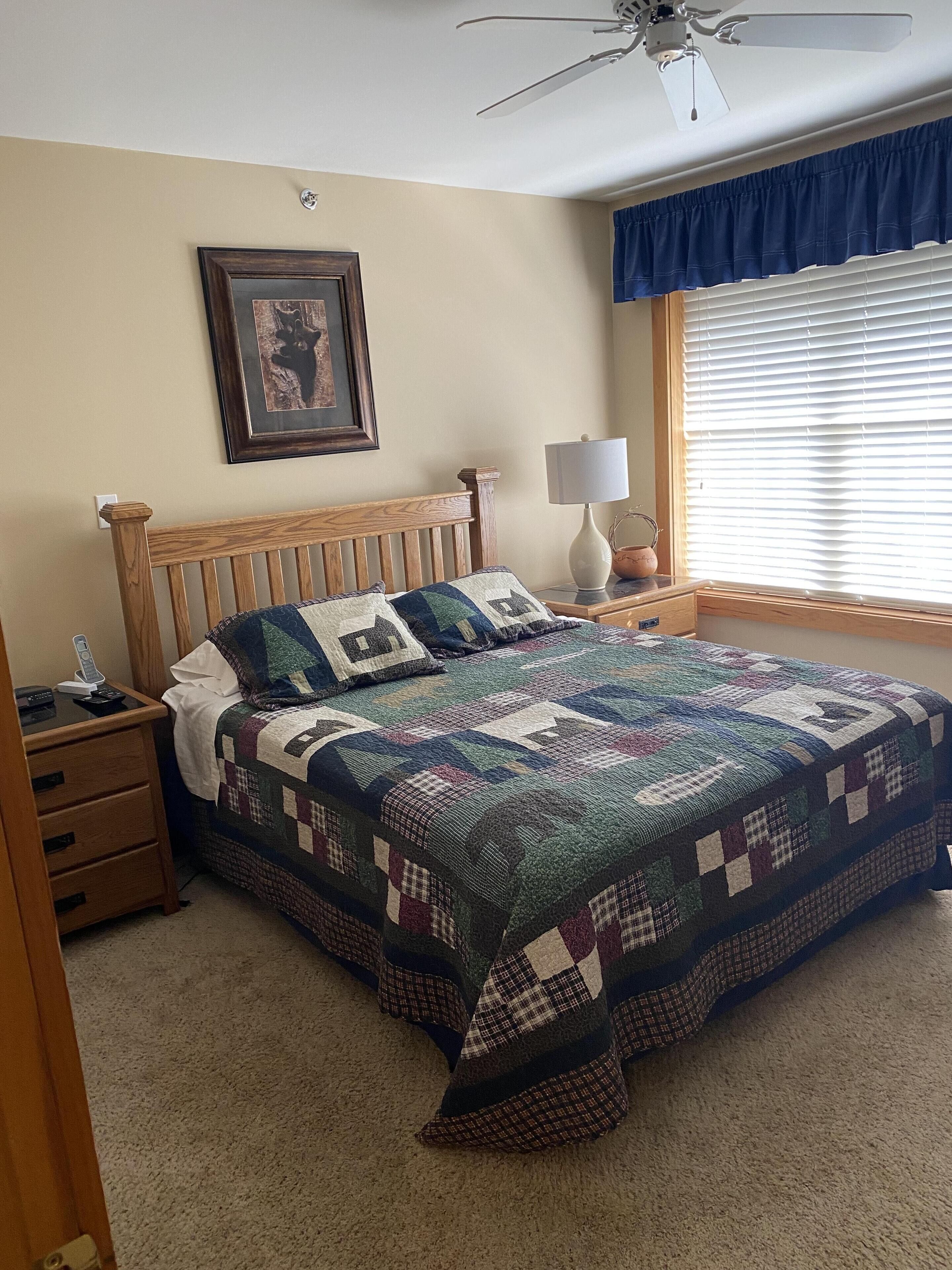 Quarto com cama de madeira, colcha, mesa de cabeceira com abajur, quadro emoldurado na parede e janela com persianas.