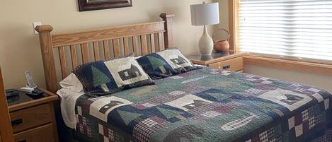 Quarto com cama de madeira, colcha, mesa de cabeceira com abajur, quadro emoldurado na parede e janela com persianas.