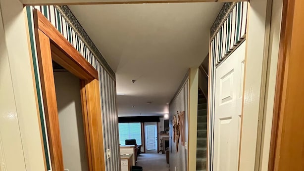 Een gang met een betegelde vloer, houten deuren en een zicht op een woonkamer met een bank en een raam.