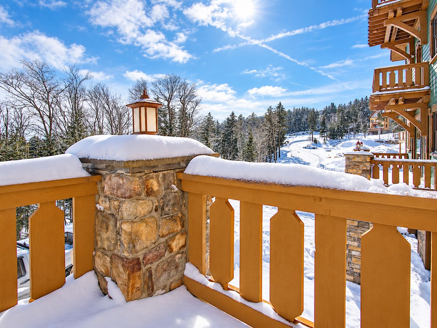 A snow-covered balcony with stone pillars, overlooking a snowy landscape and a building with a wooden balcony.