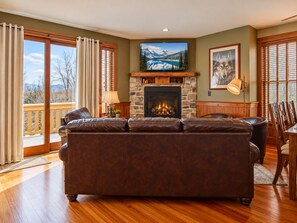 A living room with a fireplace, leather sofa, wooden flooring, and a view of a mountain lake through large windows. - Sanctuary Family Paradise Private Home With Hot tub Snowshoe Mtn Resort (Snowshoe)