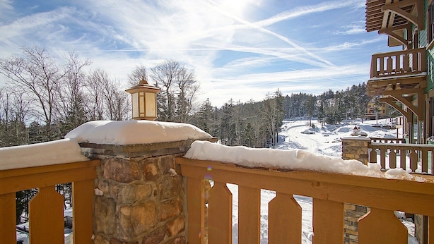 A snow-covered balcony with a stone wall and wooden railing, overlooking a snowy landscape with trees and a building in the background.