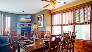 A dining area with a wooden table set for four, surrounded by chairs. A fireplace with a stone mantle is visible in the background.