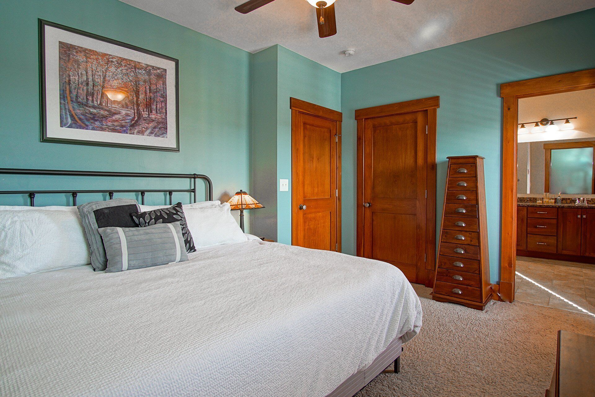 A bedroom with a bed, a nightstand, a lamp, a wooden dresser, and a framed painting on the wall.