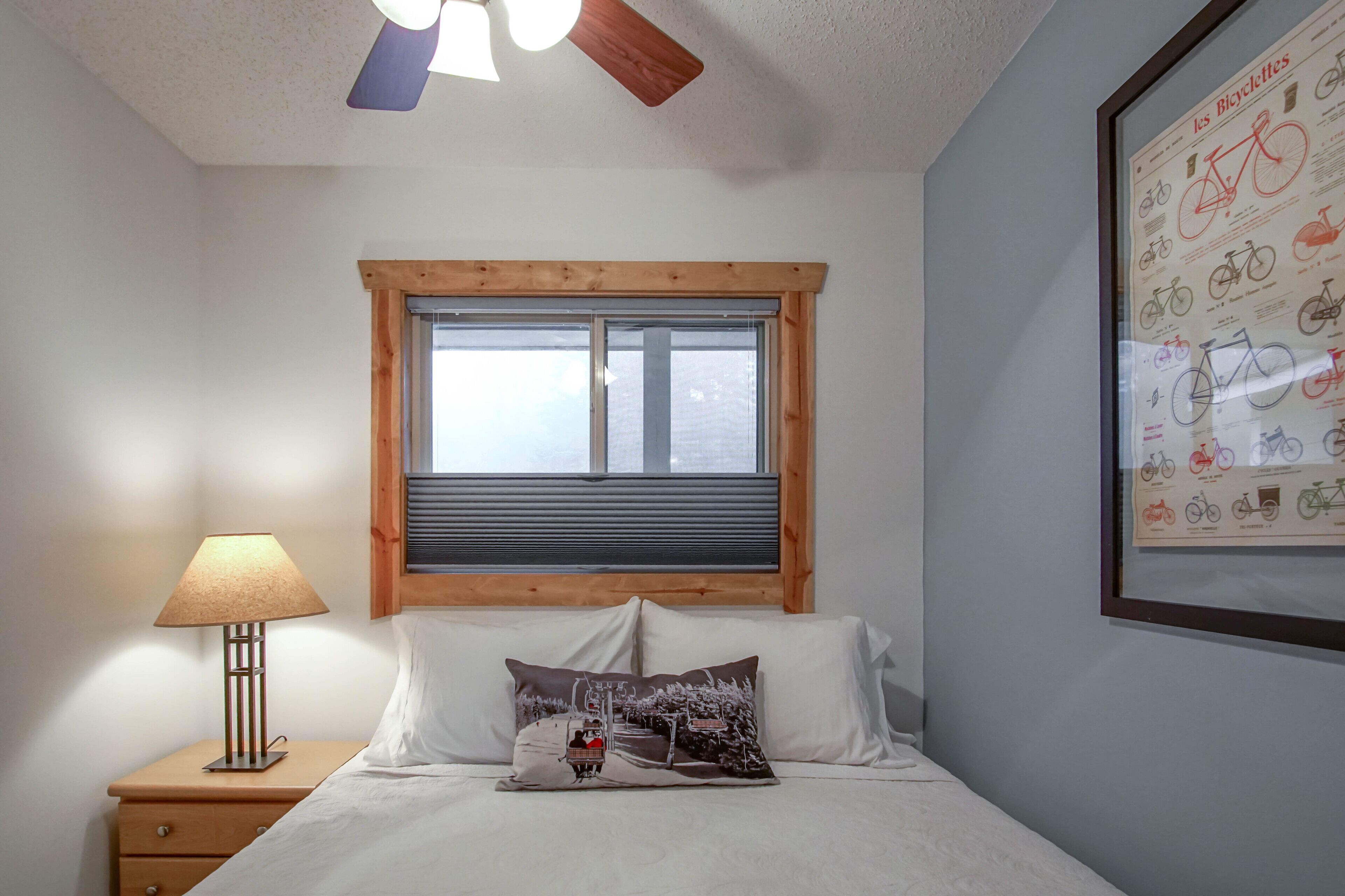 A bedroom with a wooden headboard, a window with blinds, a bedside lamp, a framed bicycle poster, and a bed with white linens.