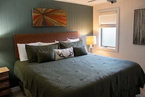 Ein Schlafzimmer mit Bett, Kissen, Kopfteil, Wandbild, Fenster mit Jalousien und einer Nachttischlampe.