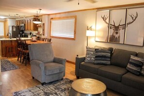 Ein Wohnzimmer mit grauem Sessel, grauem Sofa und rundem Holztisch. Es gibt einen Essbereich mit Holztisch und Stühlen. Der Raum hat Holzboden und ein großes Fenster mit Jalousien.
