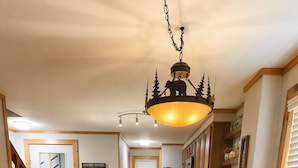 Un comedor con una mesa de madera preparada para cuatro personas, rodeada de sillas. Un candelabro cuelga encima y se ve una cocina al fondo.