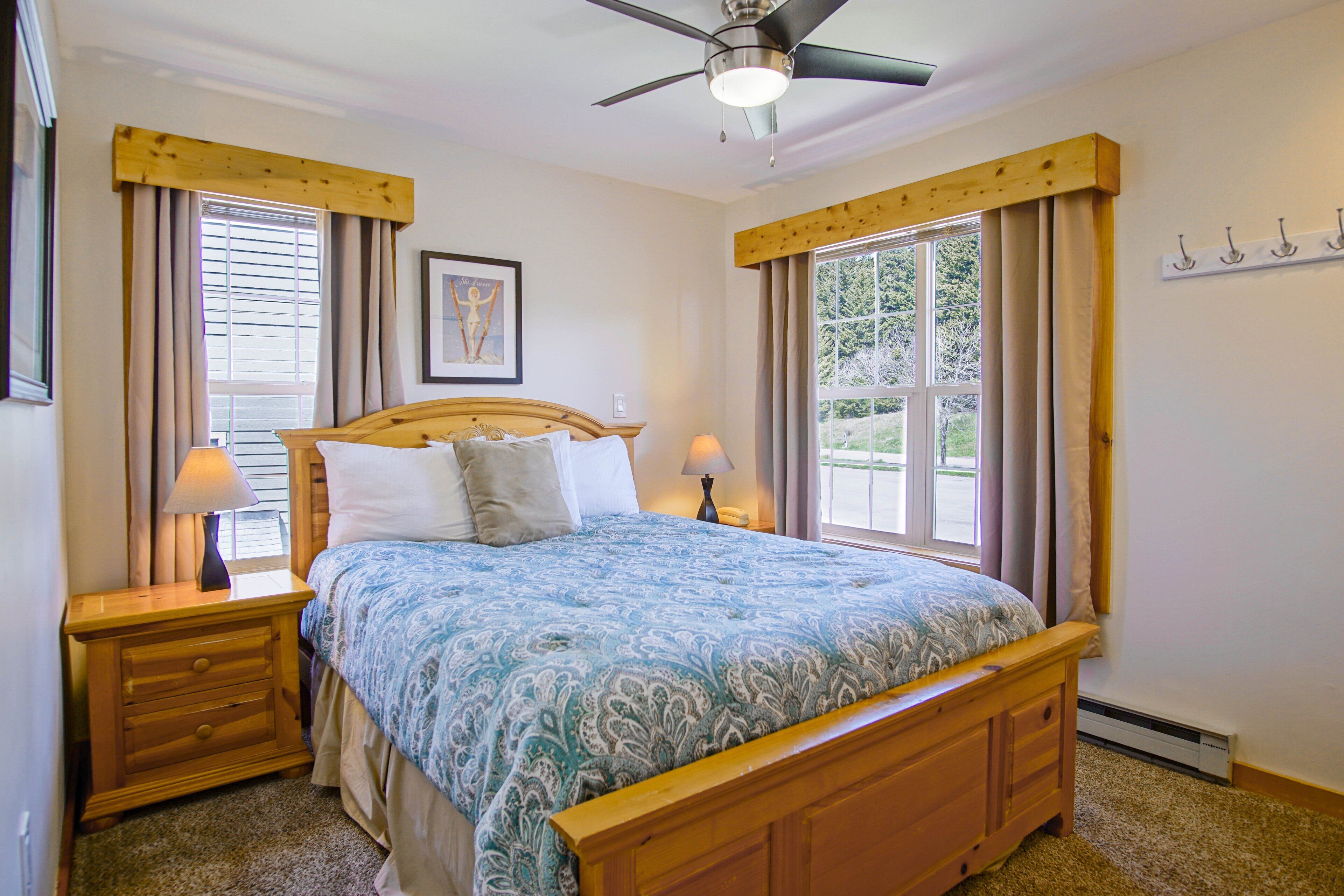 A bedroom with a wooden bed, two bedside tables, a ceiling fan, and large windows with curtains.