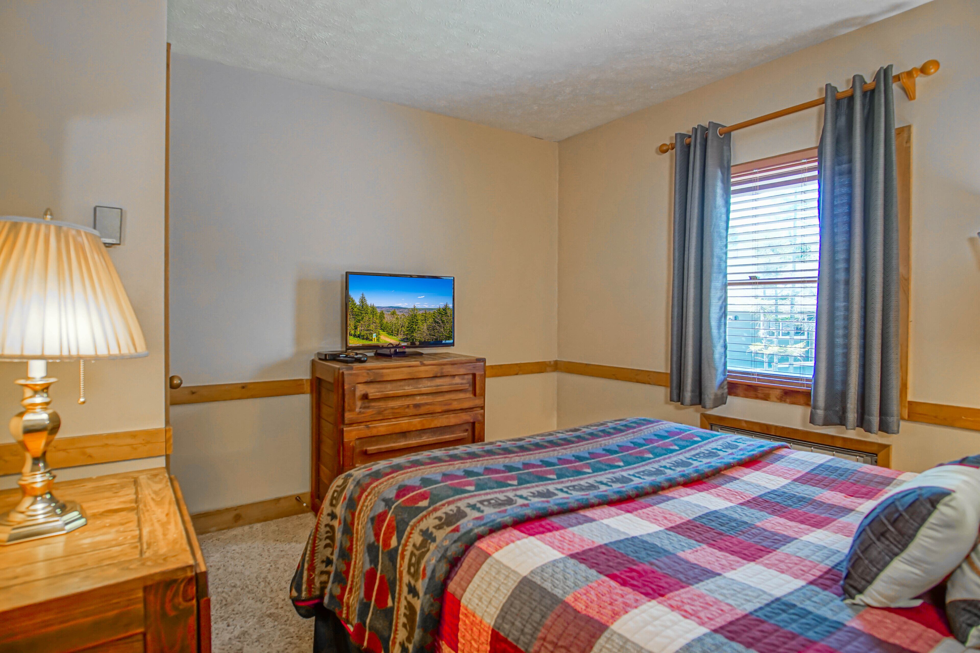 A bedroom with a wooden dresser, a television on top, a colorful quilt on the bed, and a window with curtains.