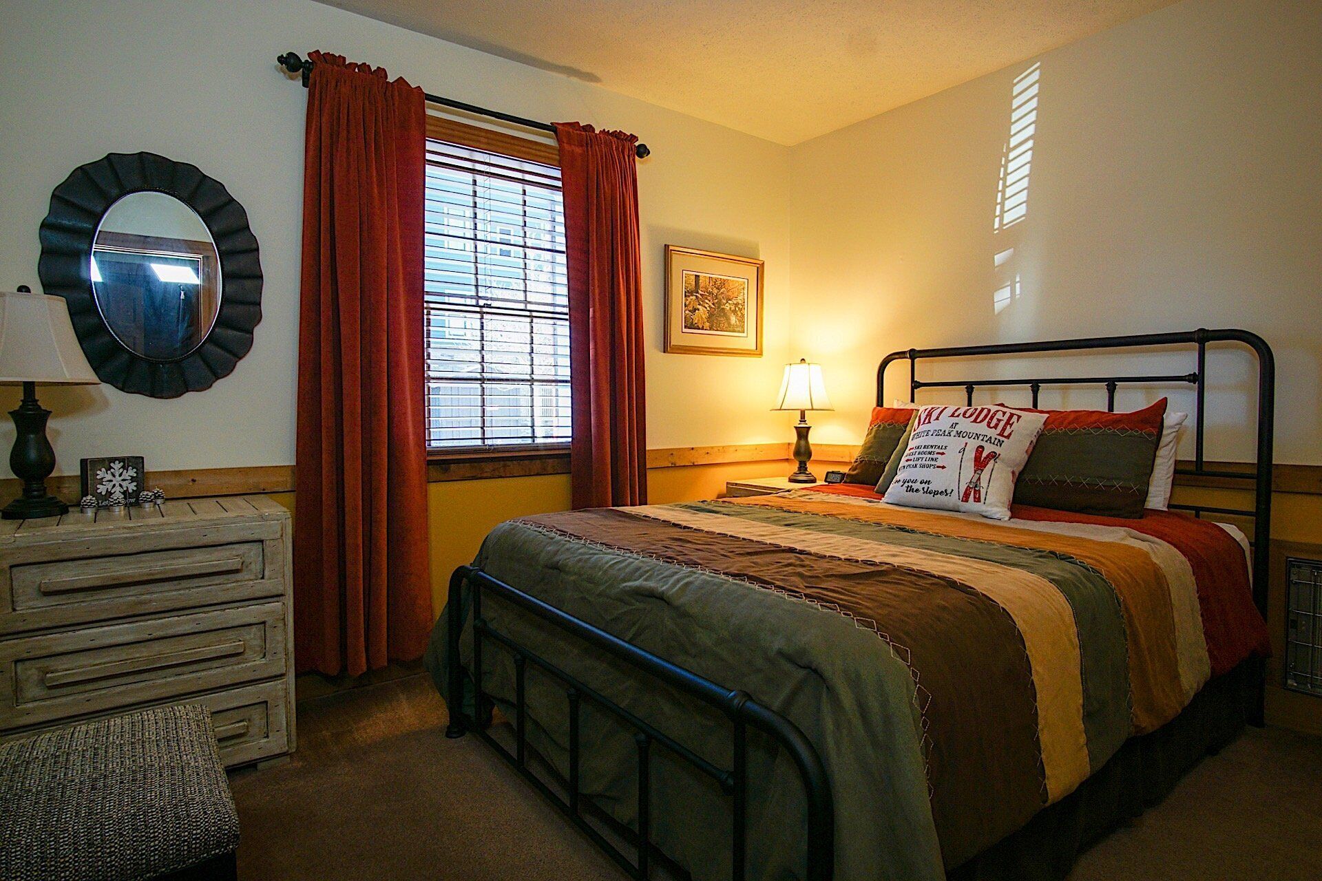 A bedroom with a bed, a dresser, a nightstand, a lamp, a window with curtains, and a framed picture on the wall.