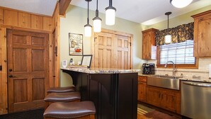 A kitchen with wooden cabinets, a bar area with stools, a coffee maker, and a window with blinds. - Whistlepunk 20 Wilderness Hideaway, Slope Side, Spacious 2 Br Condo (Snowshoe)
