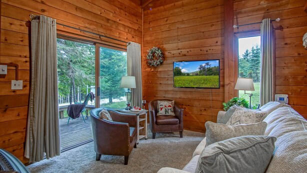 A cozy cabin interior with a wooden interior, a television mounted on the wall, a sofa, a chair, and a view of the outdoors through large windows. - Powder Monkey 4: 2 Bedrooms Plus Sleeping Loft, 2 Full Baths: Slope Side (Snowshoe)